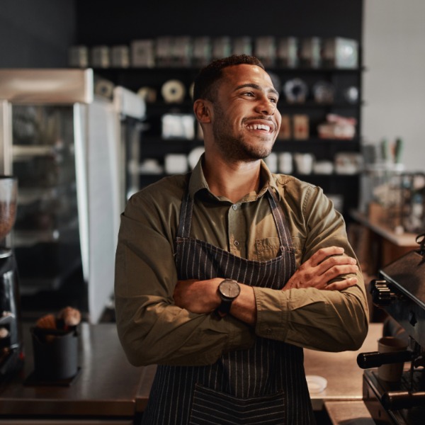 ایک مسکراتا ہوا نوجوان تہبند پہنے ایک آزاد کافی شاپ میں یسپریسو مشین پر کھڑا ہے